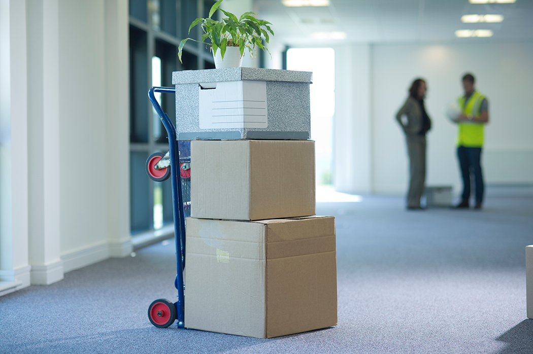 Stack of three boxes on a dolly. There is a mover and a business woman talking in the background, out of focus.